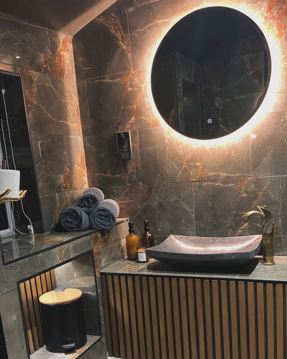 Bathroom finished in Amble: dark marble-effect tiles, a backlit round mirror and a stone basin on a slatted vanity